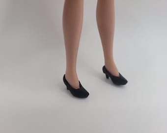Puede incluir: Un par de zapatos de muñeca negros con punta afilada y un tacón pequeño. Los zapatos se llevan puestos en un par de piernas de muñeca de piel clara que están en posición vertical. El calzado presenta un diseño sencillo en color negro sólido.