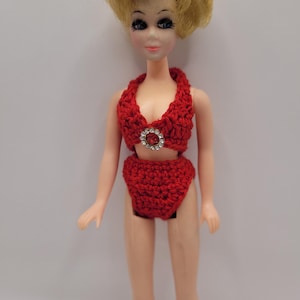 May include: A doll with blonde hair wearing a two-piece red crocheted bikini set. The top features a halter neckline with a circular silver-toned embellishment containing a central red stone surrounded by small clear crystals at the center.