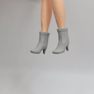 Puede incluir: Un par de botines grises con tacones peque&ntilde;os. Los botines son usados por una mu&ntilde;eca, con las piernas de la mu&ntilde;eca visibles por encima de los botines. Los botines son de color gris s&oacute;lido y tienen una apariencia texturizada.