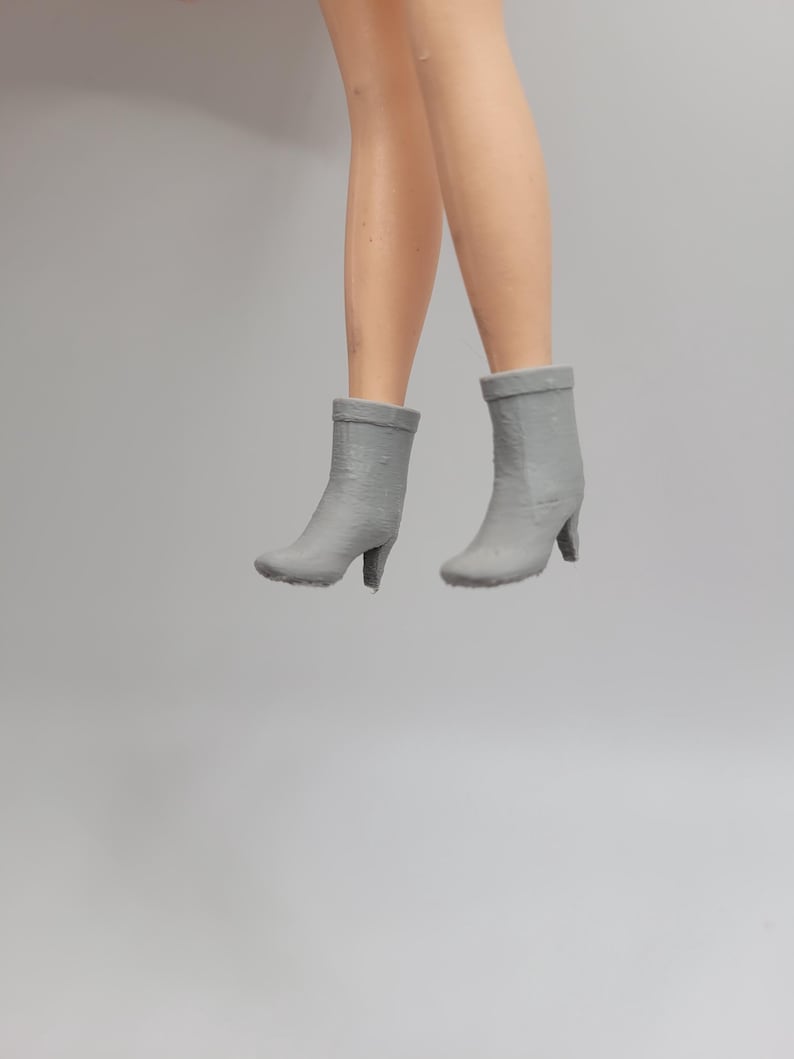 Puede incluir: Un par de botines grises con tacones peque&ntilde;os. Los botines son usados por una mu&ntilde;eca, con las piernas de la mu&ntilde;eca visibles por encima de los botines. Los botines son de color gris s&oacute;lido y tienen una apariencia texturizada.