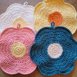 Puede incluir: Cuatro agarraderas de olla de ganchillo en forma de flor de diferentes colores. Las agarraderas de olla son blancas, amarillas, rosas y azules. Cada agarradera de olla tiene un círculo más pequeño en el centro.