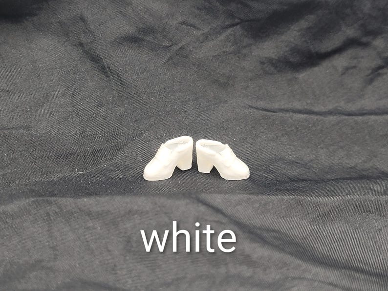 May include: A pair of miniature white shoes with a block heel and a closed-toe design. The shoes are positioned side-by-side. The word white is printed in white text at the bottom of the image, centred below the shoes.