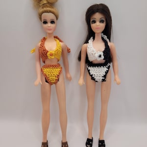 Può includere: Due bambole con tratti del viso dipinti. La bambola a sinistra ha i capelli biondi raccolti in uno chignon e indossa un bikini all'uncinetto giallo e arancione con un fiore. La bambola a destra ha lunghi capelli scuri e un bikini all'uncinetto bianco e nero.