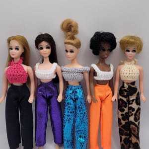 May include: Five dolls wearing different outfits. The dolls have long hair and are wearing crocheted tops and trousers. The trousers are different colours and patterns, including black, purple, blue, orange, and leopard print.