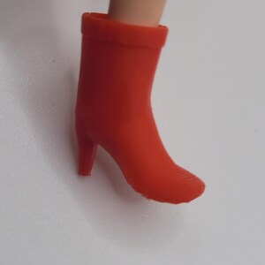 Puede incluir: Una bota de tac&oacute;n alto de color rojo brillante. La bota tiene una punta redondeada y un tac&oacute;n peque&ntilde;o. La bota est&aacute; hecha de un material liso y es de color rojo vivo s&oacute;lido. La bota es parte del atuendo de una mu&ntilde;eca.