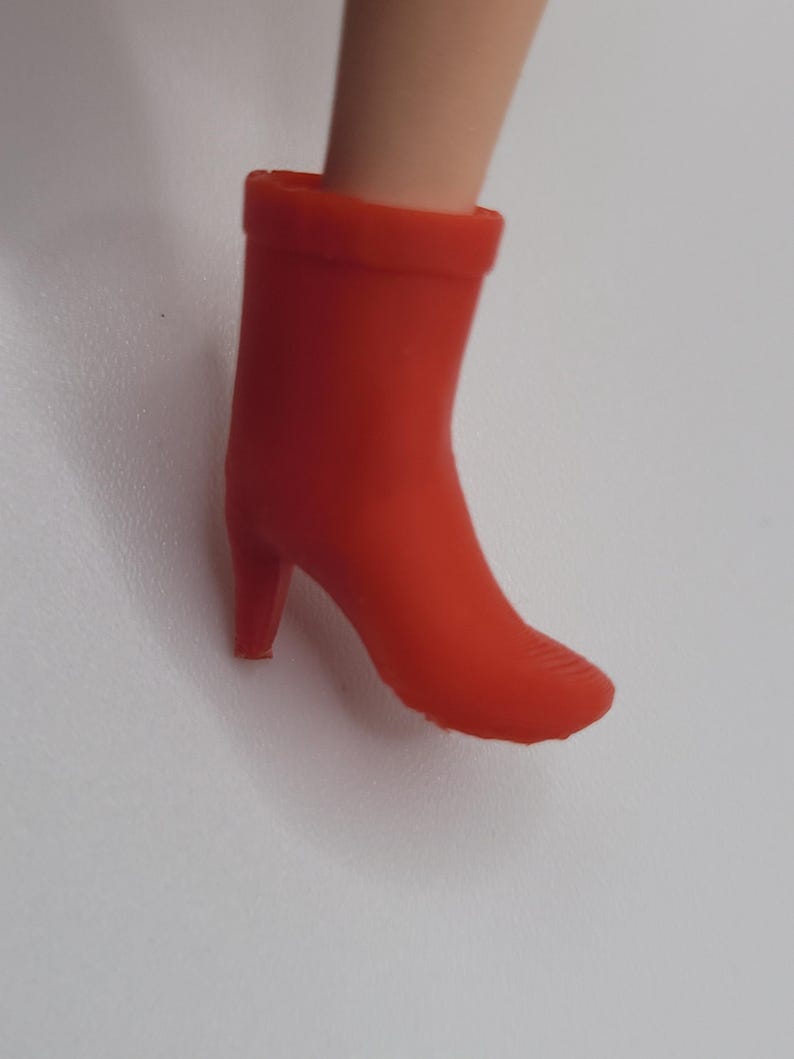 Puede incluir: Una bota de tac&oacute;n alto de color rojo brillante. La bota tiene una punta redondeada y un tac&oacute;n peque&ntilde;o. La bota est&aacute; hecha de un material liso y es de color rojo vivo s&oacute;lido. La bota es parte del atuendo de una mu&ntilde;eca.