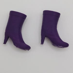 Puede incluir: Dos botas moradas en miniatura con tacones altos se muestran sobre un fondo blanco. Las botas son de color liso y parecen estar hechas de un material liso. Los tacones son delgados y puntiagudos.