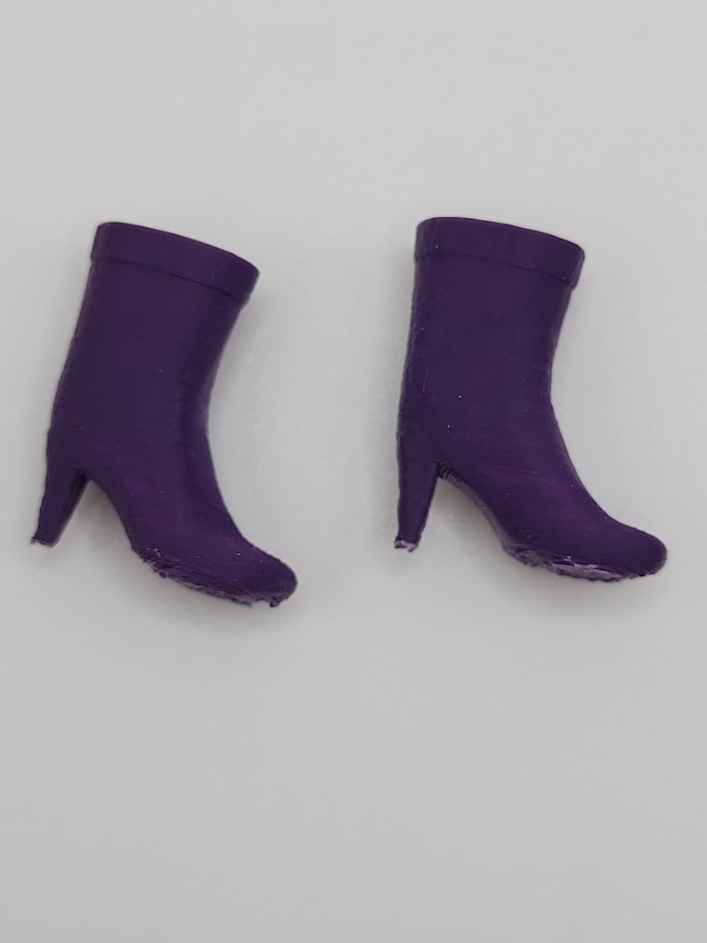 Puede incluir: Dos botas moradas en miniatura con tacones altos se muestran sobre un fondo blanco. Las botas son de color liso y parecen estar hechas de un material liso. Los tacones son delgados y puntiagudos.