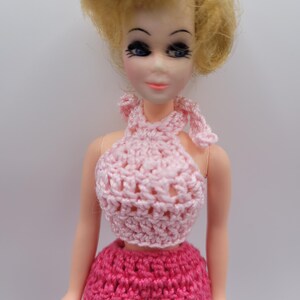 May include: A doll with blonde hair wearing a light pink crocheted halter top and dark pink crocheted pants. The doll has blue eyes, painted eyelashes, and light skin. The top ties around the neck, and the pants have a textured, woven appearance.