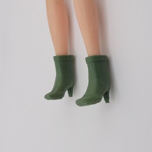 Puede incluir: Piernas de mu&ntilde;eca con botas de tac&oacute;n verde oliva. Las botas tienen una punta redondeada y un tac&oacute;n peque&ntilde;o. Las piernas son de tono claro, y las botas son el foco de la imagen.