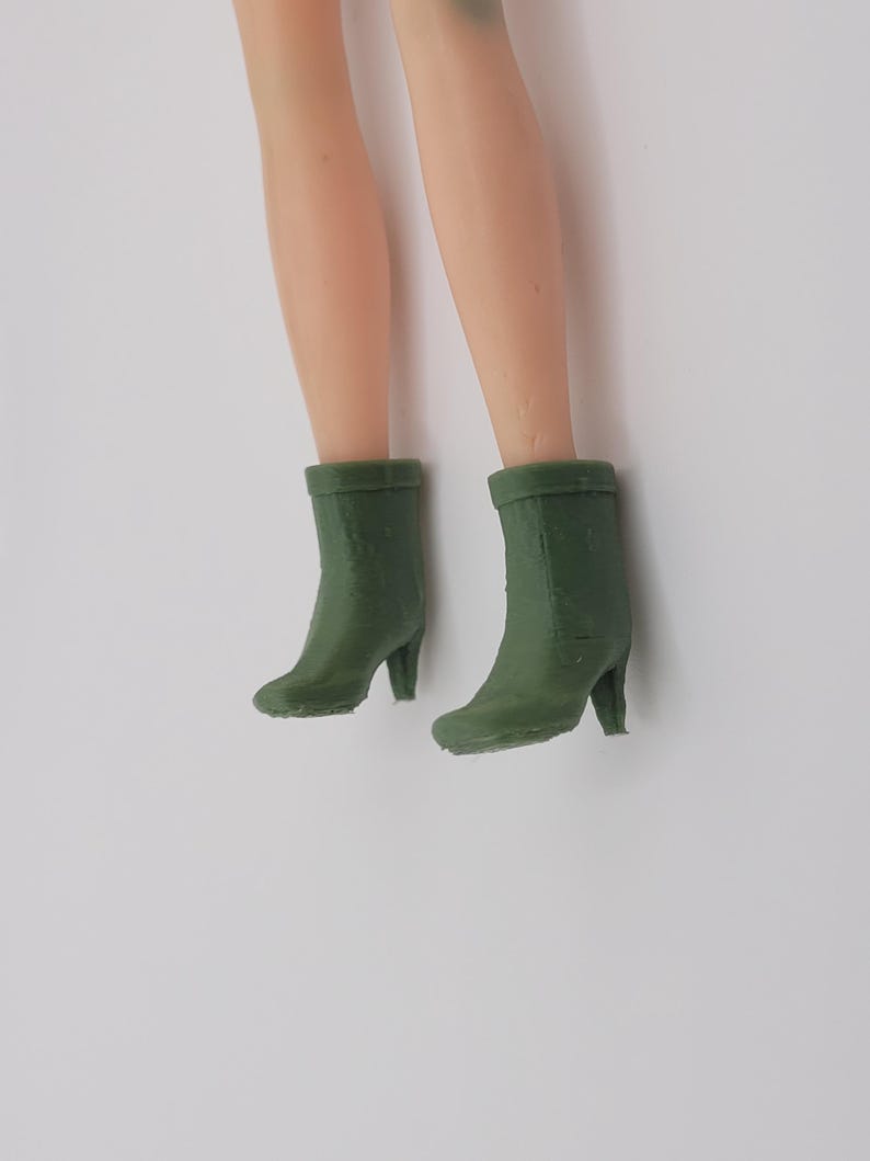 Puede incluir: Piernas de mu&ntilde;eca con botas de tac&oacute;n verde oliva. Las botas tienen una punta redondeada y un tac&oacute;n peque&ntilde;o. Las piernas son de tono claro, y las botas son el foco de la imagen.