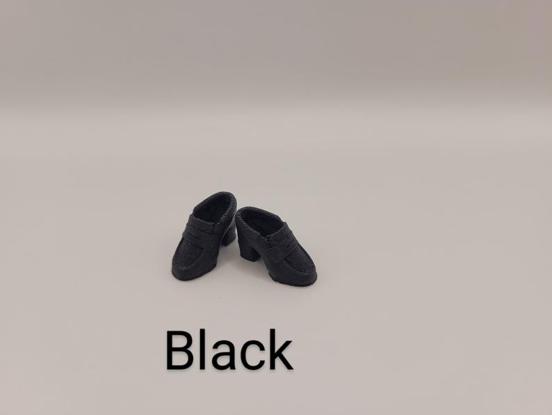 May include: A pair of miniature black loafers with a block heel and a classic penny slot strap across the vamp. The shoes are solid black in colour and feature a smooth, matte finish. The word Black is printed in large, bold, sans-serif text below.