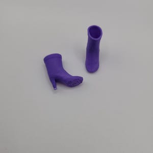 Puede incluir: Dos botas moradas de tac&oacute;n alto. Una bota est&aacute; tumbada de lado, mientras que la otra est&aacute; de pie. Las botas tienen una textura suave y una punta afilada. Los tacones son finos y delicados. El fondo es gris claro liso.
