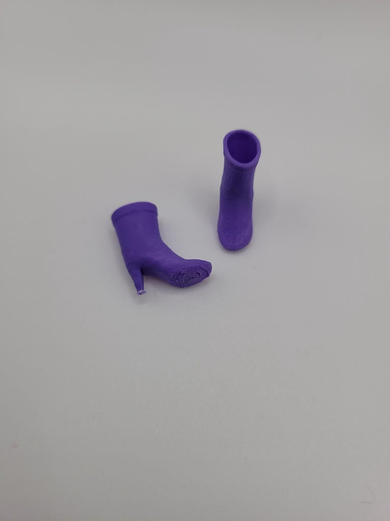 Puede incluir: Dos botas moradas de tac&oacute;n alto. Una bota est&aacute; tumbada de lado, mientras que la otra est&aacute; de pie. Las botas tienen una textura suave y una punta afilada. Los tacones son finos y delicados. El fondo es gris claro liso.