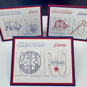 May include: Three white cards with the word "HERO" in large, bold, red outlined letters. Each card features a different symbol representing a first responder: a police officer's hat, a firefighter's helmet, and a medical star of life. Each card includes the text "a HERO is an ordinary person who faces extraordinary circumstances with COURAGE, HONOR and SELF-SACRIFICE" and "with gratitude".
