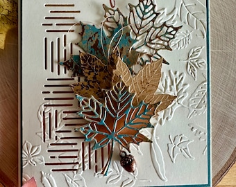 May include: A greeting card with the text "TRULY Thankful" in brown script. The card features a white embossed background with leaf patterns, a vertical arrangement of die-cut maple leaves in blue and bronze tones, and a small copper acorn charm.