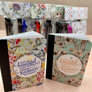 May include: Two small notebooks with floral covers and black spines. The left notebook has a white circular label with blue text reading "HOW BEAUTIFUL A DAY CAN BE WHEN KINDNESS TOUCHES IT." The right notebook has a similar label with brown text.