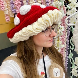 May include: A red and white crocheted mushroom hat with white polka dots. The hat is worn by a person with long brown hair and glasses.