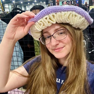 May include: A person wearing a purple and cream crocheted hat with a ruffled brim. The hat is worn over long brown hair and the person is wearing glasses.