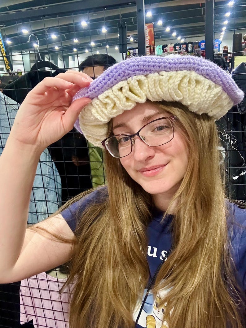 May include: A person wearing a purple and cream crocheted hat with a ruffled brim. The hat is worn over long brown hair and the person is wearing glasses.