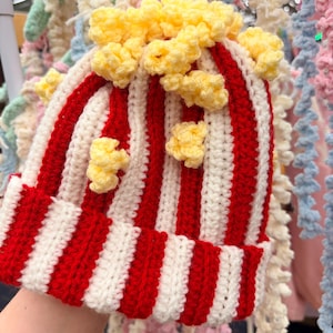 May include: A handmade, crocheted beanie hat with a popcorn design. The hat has red and white stripes, with yellow popcorn details. A quirky and unique accessory.