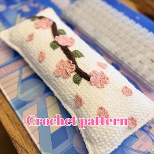 May include: A white crochet pillow with a floral design. The pillow features a brown branch with pink flowers and green leaves. Scattered pink petals add detail. The text "Crochet pattern" is displayed on the pillow.
