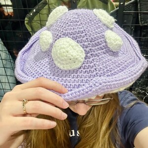 May include: A purple crocheted hat with white crocheted circles. The hat is being worn by a person with long brown hair.