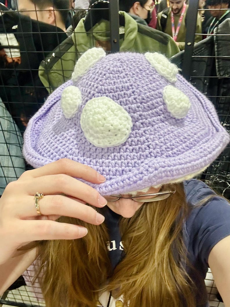 May include: A purple crocheted hat with white crocheted circles. The hat is being worn by a person with long brown hair.