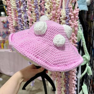 May include: A pink crocheted hat with white crocheted dots. The hat is being held by a hand.