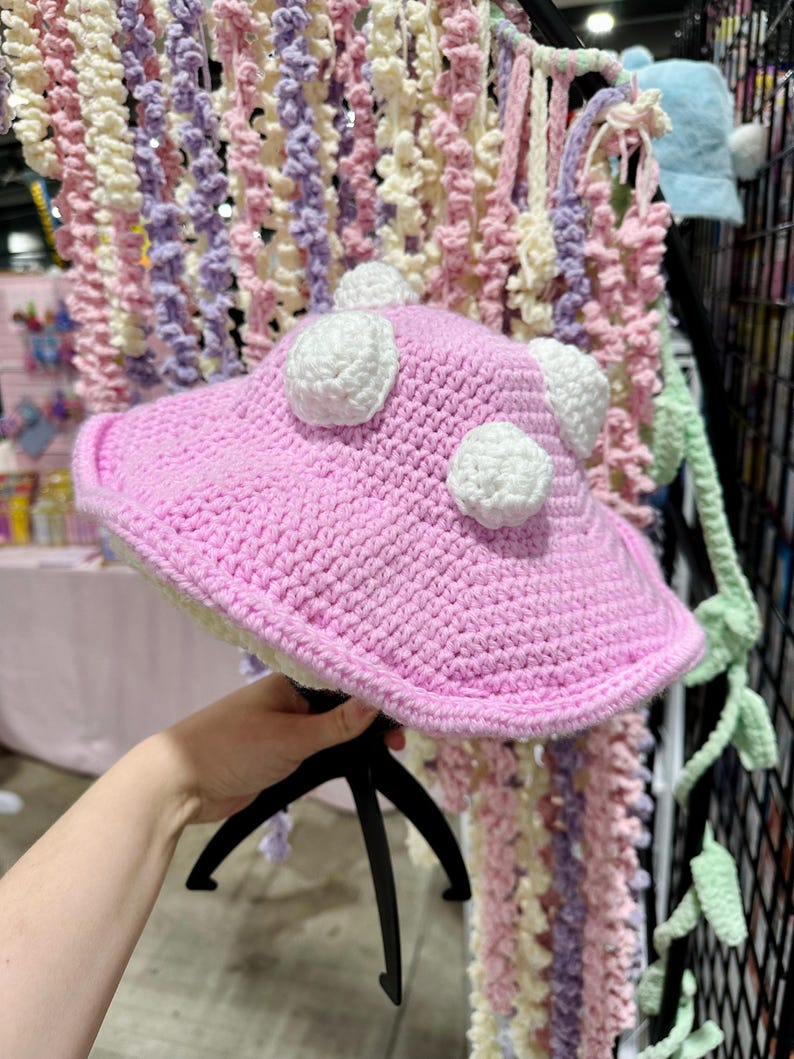 May include: A pink crocheted hat with white crocheted dots. The hat is being held by a hand.