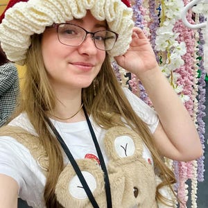 May include: A woman wearing a red and white crocheted hat with a white crocheted bear overalls. The hat has a ruffled edge and the overalls have a bear face on the front.