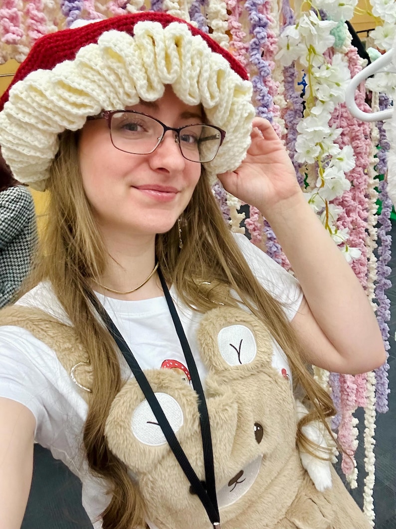 May include: A woman wearing a red and white crocheted hat with a white crocheted bear overalls. The hat has a ruffled edge and the overalls have a bear face on the front.