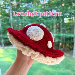 May include: A red and white crocheted mushroom hat with a ruffled cream-coloured brim and white circular spots on the cap. The text "Crochet pattern" is displayed in pink letters above the hat, which features a textured yarn construction.