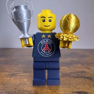 Puede incluir: Una minifigura de pl&aacute;stico amarilla con un torso azul marino que muestra el logotipo del Paris Saint-Germain y dos estrellas doradas. La figura sostiene un trofeo plateado en la mano derecha y un trofeo de bal&oacute;n de f&uacute;tbol dorado en la izquierda.