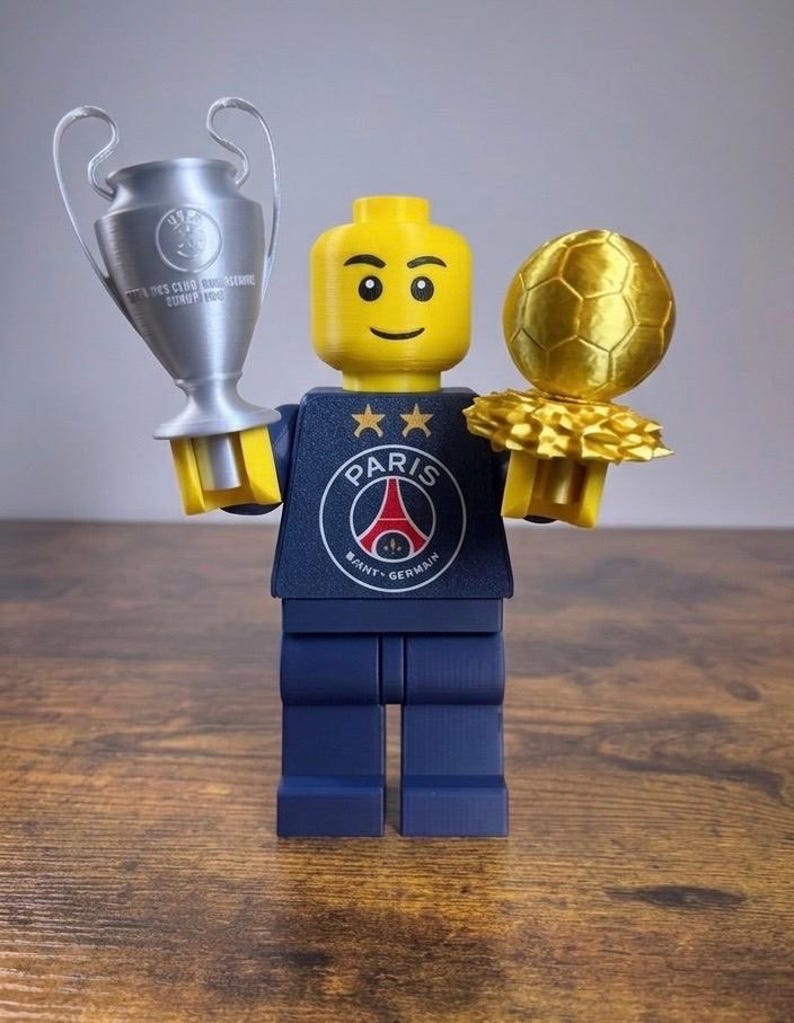 Puede incluir: Una minifigura de pl&aacute;stico amarilla con un torso azul marino que muestra el logotipo del Paris Saint-Germain y dos estrellas doradas. La figura sostiene un trofeo plateado en la mano derecha y un trofeo de bal&oacute;n de f&uacute;tbol dorado en la izquierda.
