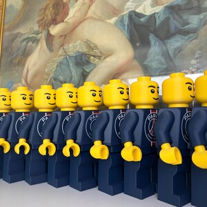 Puede incluir: Una fila de figuras de juguete de pl&aacute;stico id&eacute;nticas con cabezas y manos amarillas, vistiendo torsos y piernas azul oscuro. Cada torso presenta un logotipo circular del Paris Saint-Germain con el texto PARIS SAINT-GERMAIN en blanco.