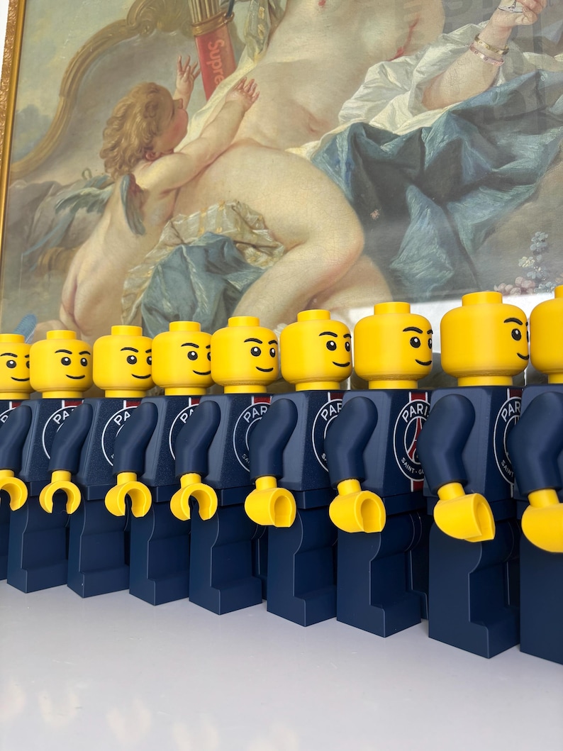 Puede incluir: Una fila de figuras de juguete de pl&aacute;stico id&eacute;nticas con cabezas y manos amarillas, vistiendo torsos y piernas azul oscuro. Cada torso presenta un logotipo circular del Paris Saint-Germain con el texto PARIS SAINT-GERMAIN en blanco.