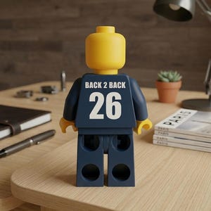 Puede incluir: Una figura de juguete de pl&aacute;stico amarillo con una cabeza lisa y redondeada y un torso azul oscuro. La parte posterior del torso presenta el texto blanco BACK 2 BACK sobre el n&uacute;mero 26 en blanco grande. La figura tiene brazos y piernas articulados.