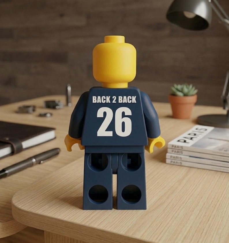 Puede incluir: Una figura de juguete de pl&aacute;stico amarillo con una cabeza lisa y redondeada y un torso azul oscuro. La parte posterior del torso presenta el texto blanco BACK 2 BACK sobre el n&uacute;mero 26 en blanco grande. La figura tiene brazos y piernas articulados.
