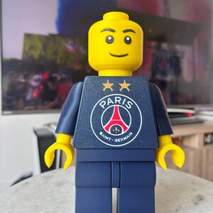 Puede incluir: Una figura de pl&aacute;stico amarilla grande con torso y piernas de color azul oscuro. El torso presenta el logotipo del club de f&uacute;tbol Paris Saint-Germain en blanco, rojo y azul, coronado por dos estrellas doradas. Tiene una cara sonriente simple.