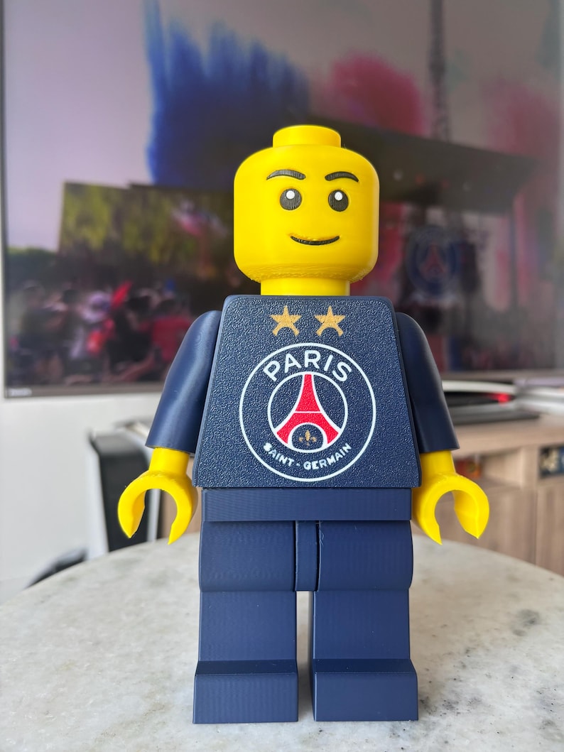 Puede incluir: Una figura de pl&aacute;stico amarilla grande con torso y piernas de color azul oscuro. El torso presenta el logotipo del club de f&uacute;tbol Paris Saint-Germain en blanco, rojo y azul, coronado por dos estrellas doradas. Tiene una cara sonriente simple.