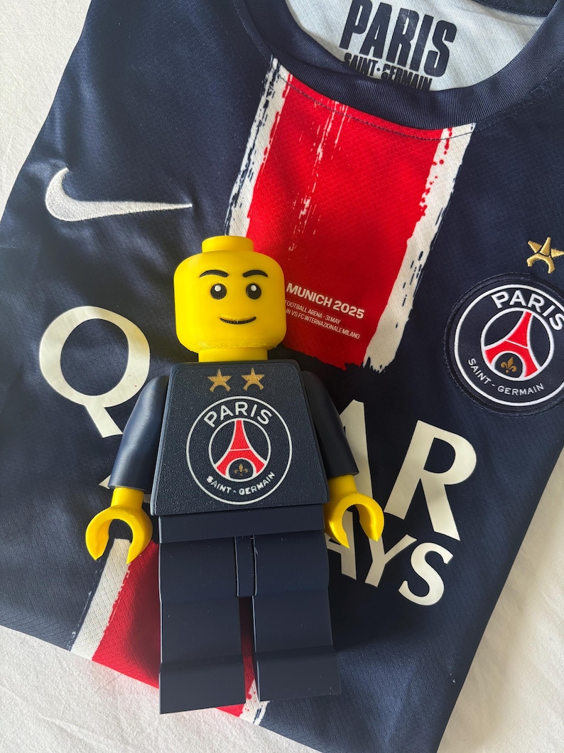 Puede incluir: Una camiseta de f&uacute;tbol azul oscuro del Paris Saint-Germain con una franja vertical roja central, un logo de Nike blanco y el texto MUNICH 2025. Una minifigura de pl&aacute;stico amarilla con torso azul oscuro y el logo del Paris Saint-Germain est&aacute; encima.