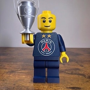 Puede incluir: Una minifigura de pl&aacute;stico amarilla que viste un torso azul oscuro con el logotipo del Paris Saint-Germain y dos estrellas doradas encima. La figura tiene manos amarillas y sostiene un trofeo plateado con dos asas en su mano derecha.