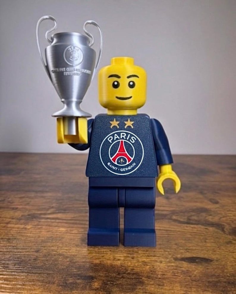 Puede incluir: Una minifigura de pl&aacute;stico amarilla que viste un torso azul oscuro con el logotipo del Paris Saint-Germain y dos estrellas doradas encima. La figura tiene manos amarillas y sostiene un trofeo plateado con dos asas en su mano derecha.