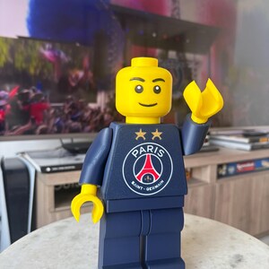 Puede incluir: Una figura de juguete de pl&aacute;stico con cabeza y manos amarillas, que viste un torso y piernas de color azul oscuro. El torso presenta el logotipo del club de f&uacute;tbol Paris Saint-Germain en blanco, rojo y azul, con dos estrellas doradas.