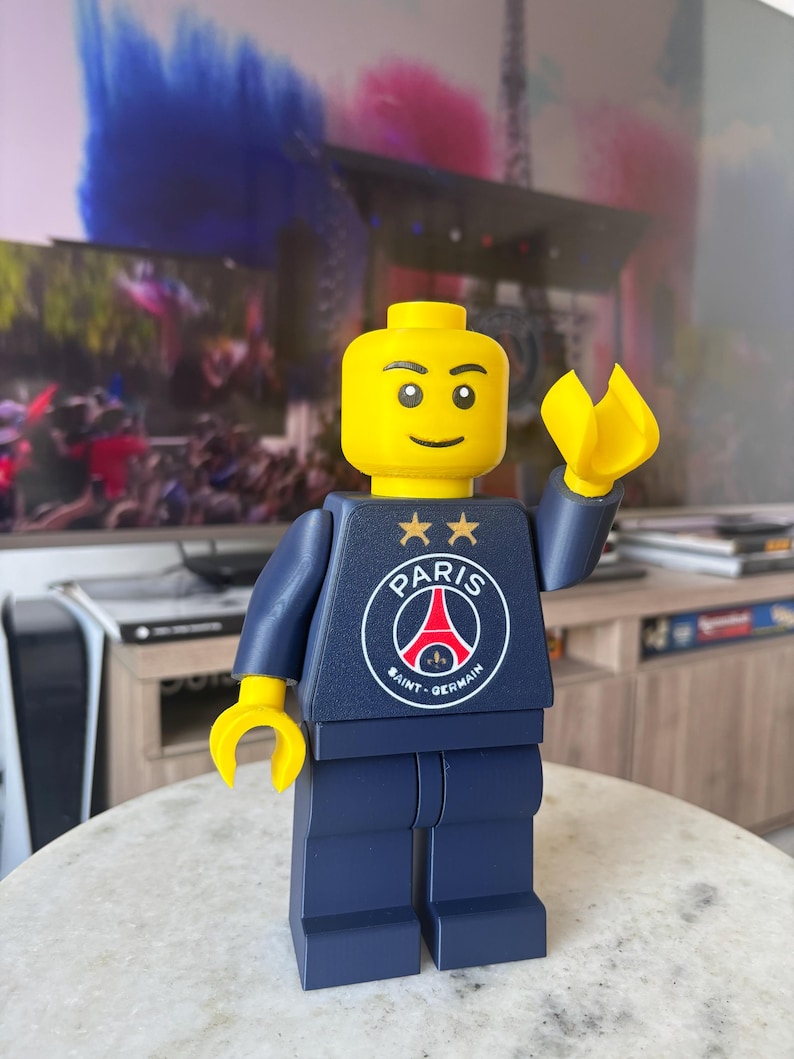 Puede incluir: Una figura de juguete de pl&aacute;stico con cabeza y manos amarillas, que viste un torso y piernas de color azul oscuro. El torso presenta el logotipo del club de f&uacute;tbol Paris Saint-Germain en blanco, rojo y azul, con dos estrellas doradas.