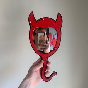 May include: A red devil-shaped hand mirror with black outlines. The mirror is held by a hand in front of a white wall.