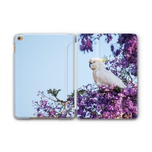 May include: An open tablet case featuring a white cockatoo perched in a tree with purple flowers. The left side of the case is light blue. The cockatoo has a yellow crest and is surrounded by purple blossoms.