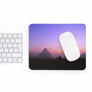 以下が含まれることがあります： 砂漠に3つのピラミッドがそびえ立つ、紫とピンクの夕焼け。マウスパッドの上に白いコンピューターマウスが置かれている。