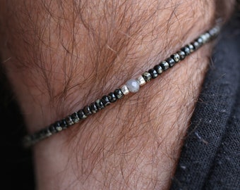Pode incluir: Uma pulseira de contas com pequenas contas escuras com um padrão salpicado. Uma única conta cinza claro e contas prateadas são centralizadas na pulseira. A pulseira é usada no pulso.
